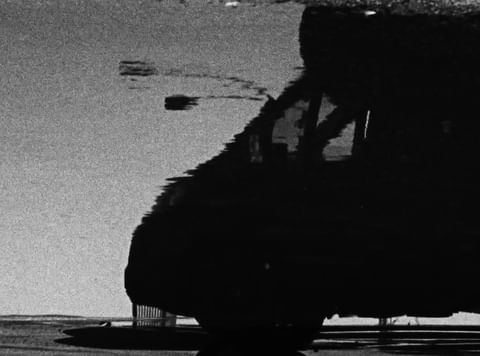 Abstract black and white image featuring the silhouette of a vehicle reflected in a puddle on a wet ground surface.