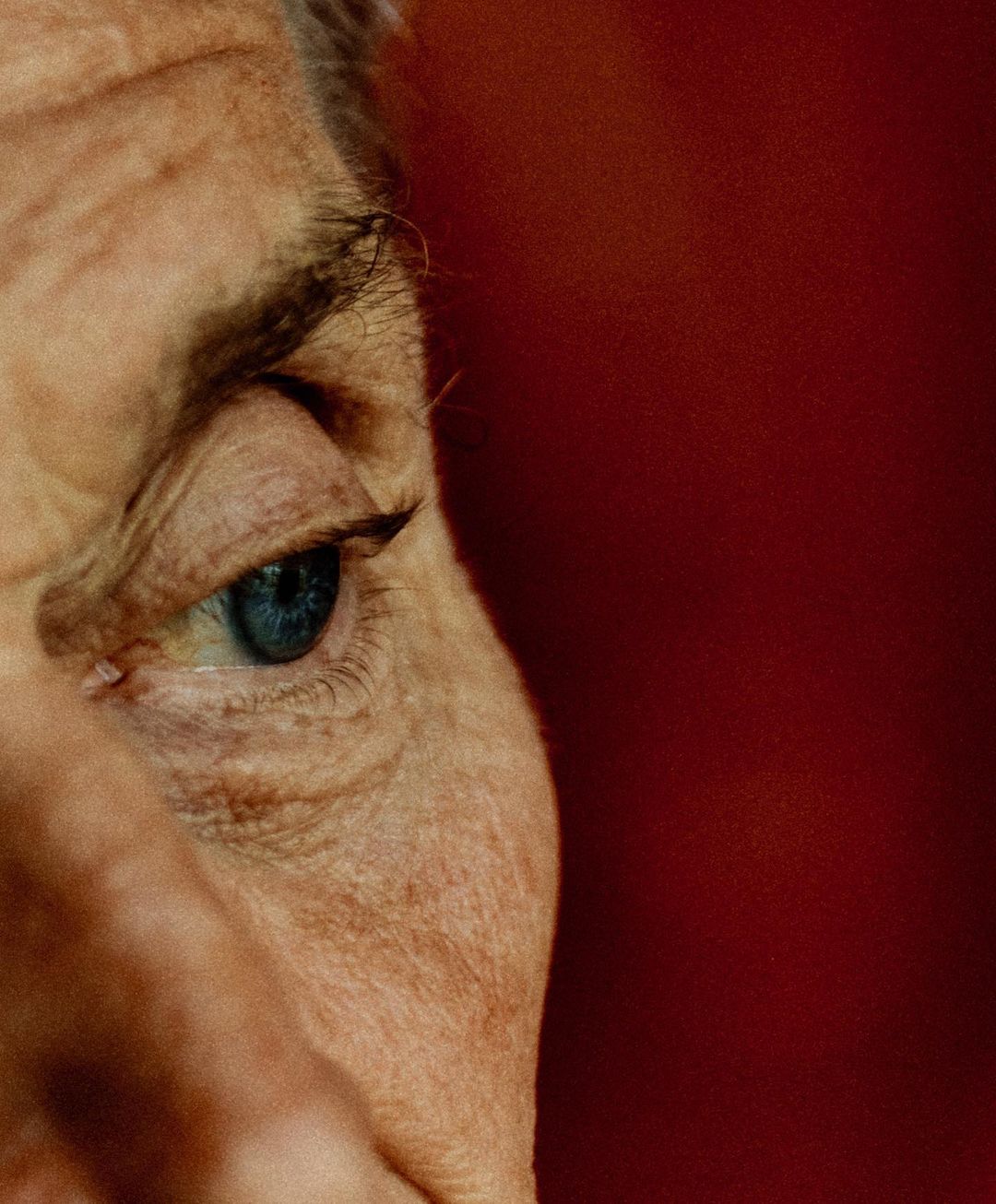 Close-up of an adult's face, showcasing a blue eye and surrounding skin texture against a deep red background. Focus on detail.