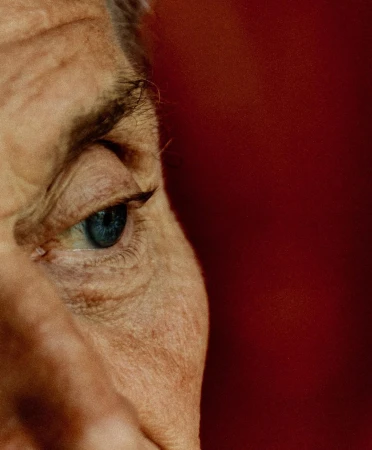 Close-up of an adult's face, showcasing a blue eye and surrounding skin texture against a deep red background. Focus on detail.