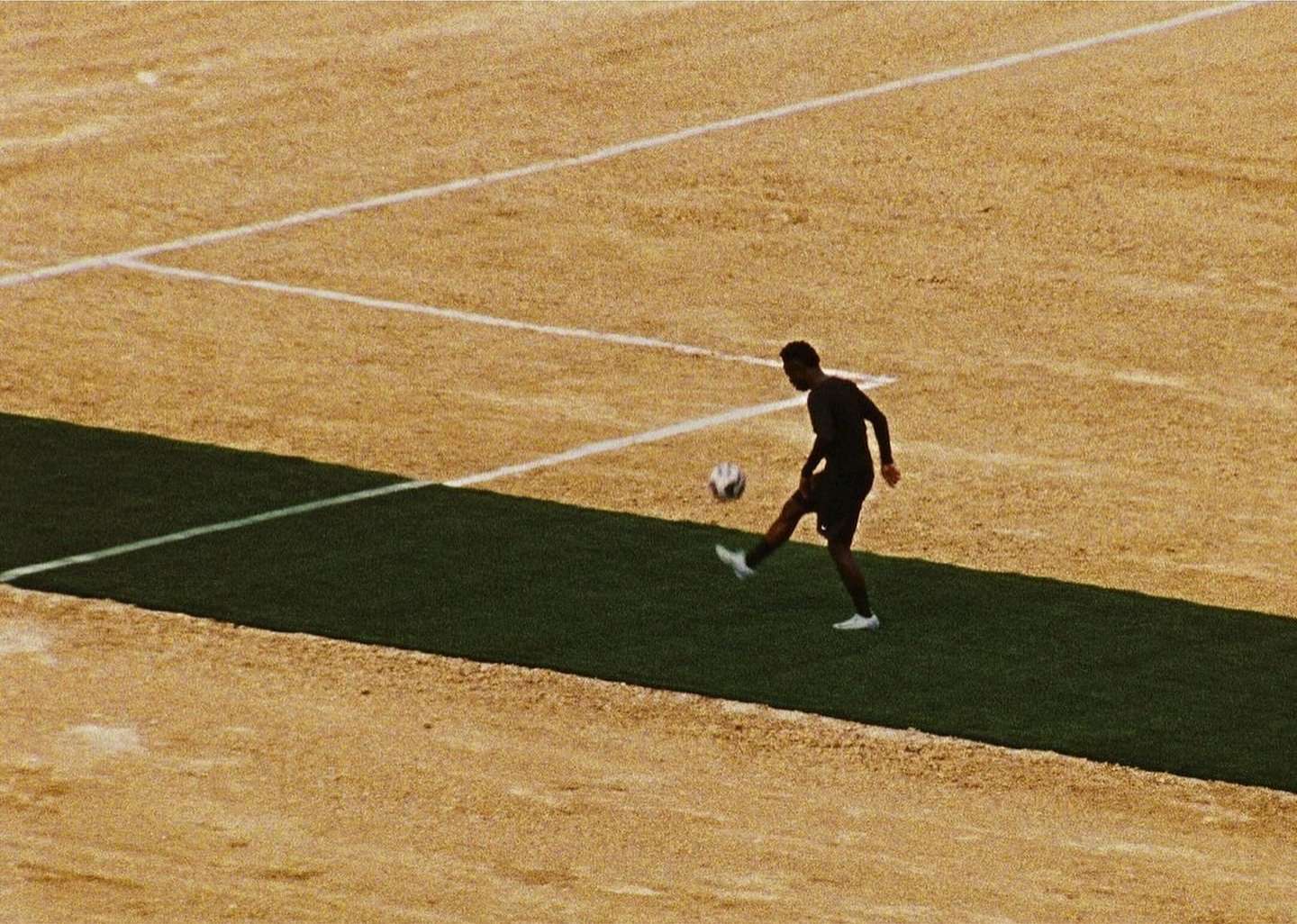 Young man in sports clothing kicks a soccer ball in a sunlit outdoor training area with a brown and green field in the background.