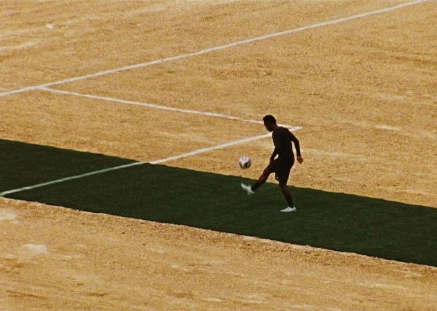 Young man in sports clothing kicks a soccer ball in a sunlit outdoor training area with a brown and green field in the background.