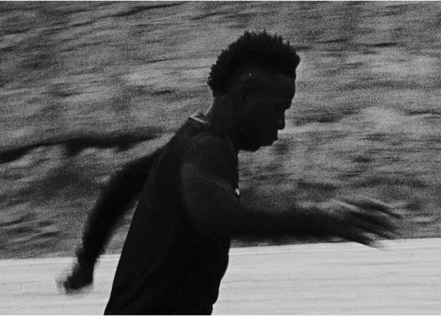 Action shot of a Black male athlete with a mohawk running outdoors in black and white, capturing motion and focus on speed.