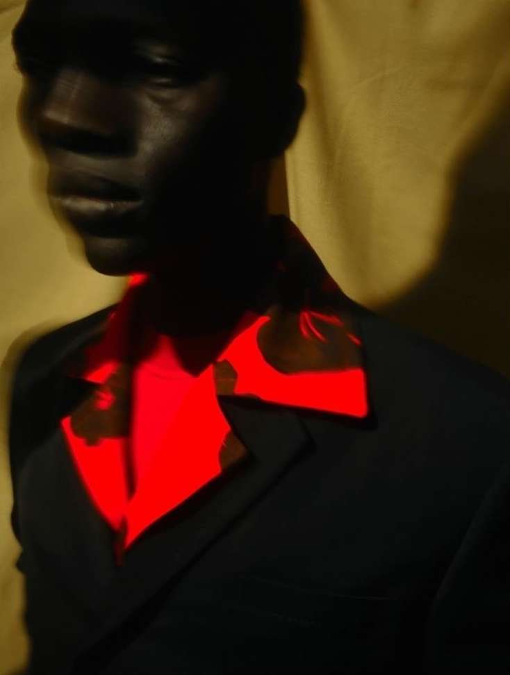 Portrait of a man with dark skin wearing a black suit and red patterned shirt against a golden fabric backdrop in a studio setting.