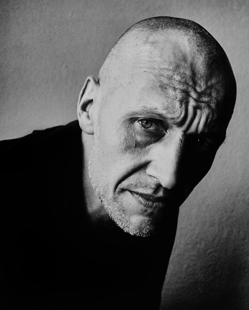 Portrait of a bald adult man with light stubble in a black shirt, looking intensely at the camera against a gray background in black and white.