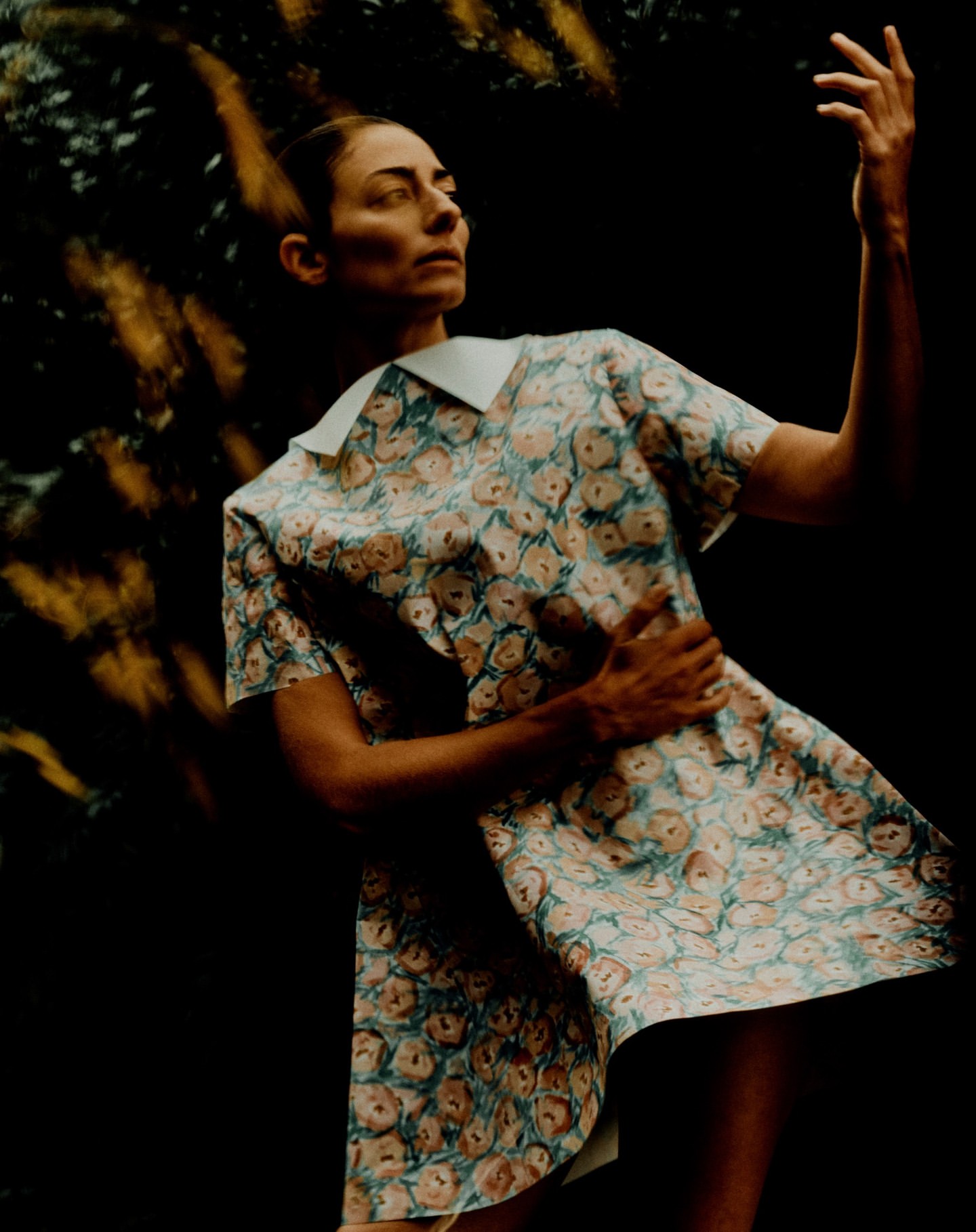 Woman in a floral dress stands with a raised hand against a dark background with blurred golden light accents, captured in a medium shot.
