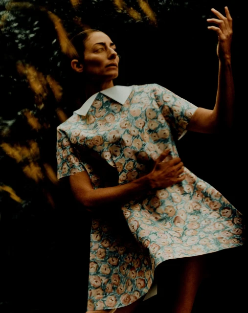Woman in a floral dress stands with a raised hand against a dark background with blurred golden light accents, captured in a medium shot.