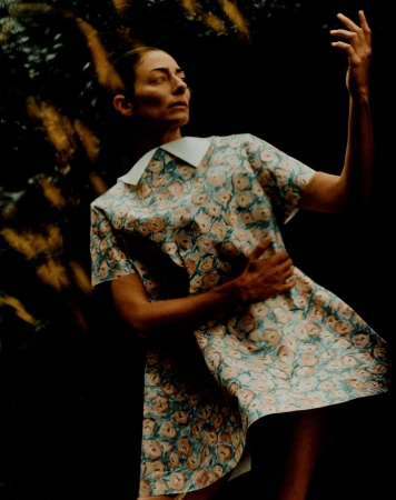 Woman in a floral dress stands with a raised hand against a dark background with blurred golden light accents, captured in a medium shot.