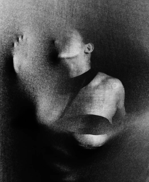Artistic black and white portrait of a person with head and shoulders shrouded in textured fabric against a dark, shadowy backdrop.