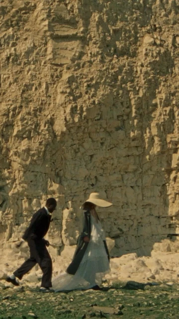 An adult male in a suit and an adult female in a long white dress and hat walk in front of a light brown cliff face.