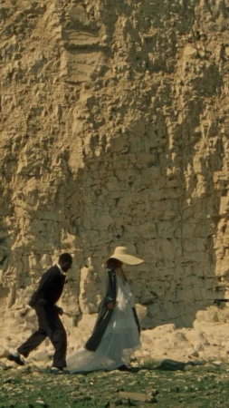 An adult male in a suit and an adult female in a long white dress and hat walk in front of a light brown cliff face.