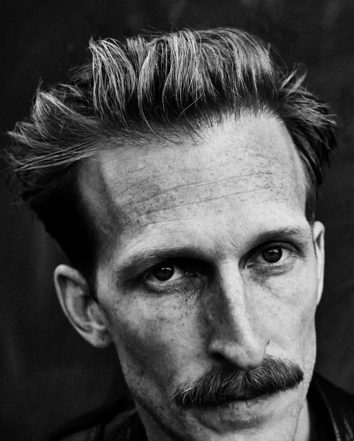 Black and white portrait of a fair-skinned adult male with a mustache, looking directly at the camera, against a dark background.