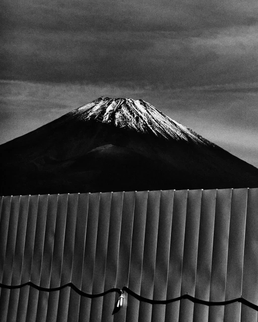 Monochrome image of Mount Fuji with snow-capped peak, dark cloudy sky, and corrugated metal fence in foreground on an overcast day.