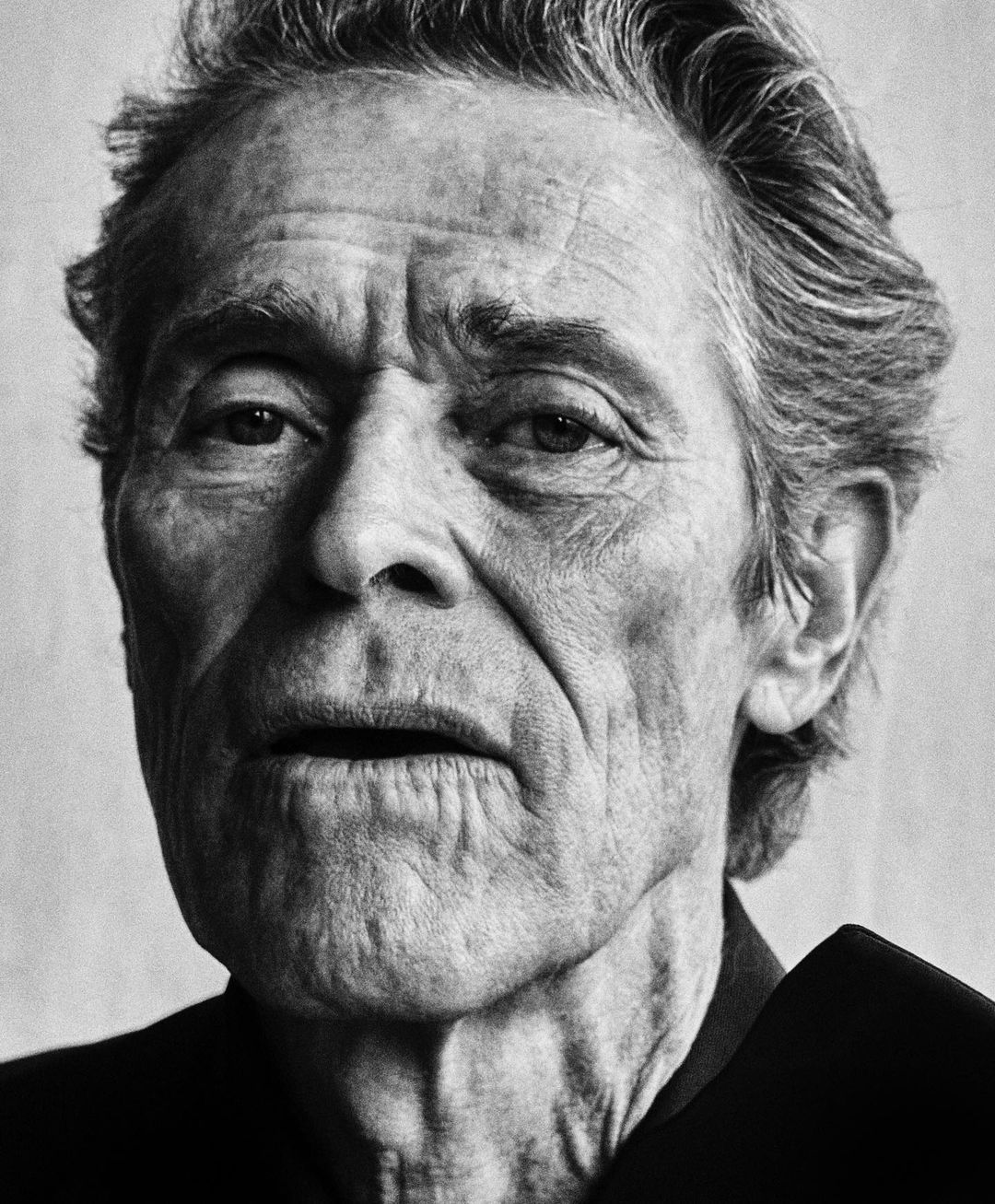 Black and white portrait of an elderly man with wrinkles, short gray hair, wearing a dark shirt, against a neutral background.