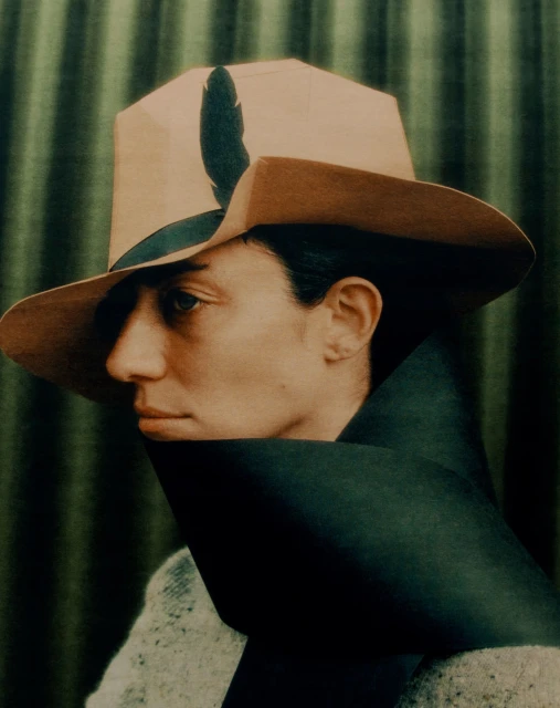 Profile portrait of an adult wearing a beige hat with a dark feather and a black scarf in front of a green striped background.