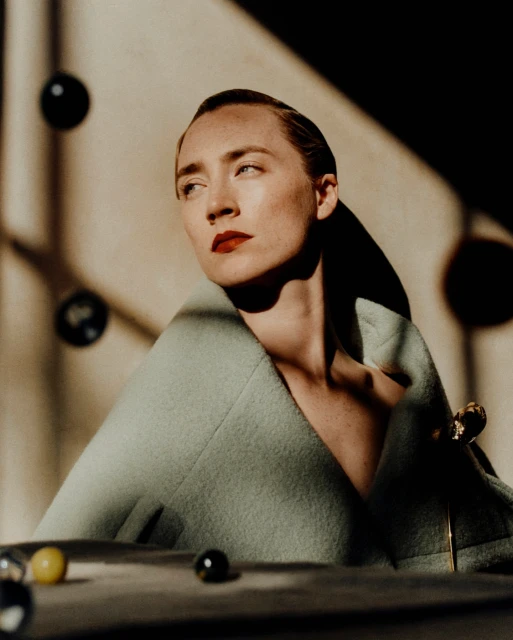 Portrait of a blonde female model with red lipstick, wearing a green coat in a studio with spheres, shadows and props.
