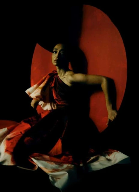 Dramatic studio portrait of an Asian female in a dark red velvet gown with a white floral hem, posing against a black background.