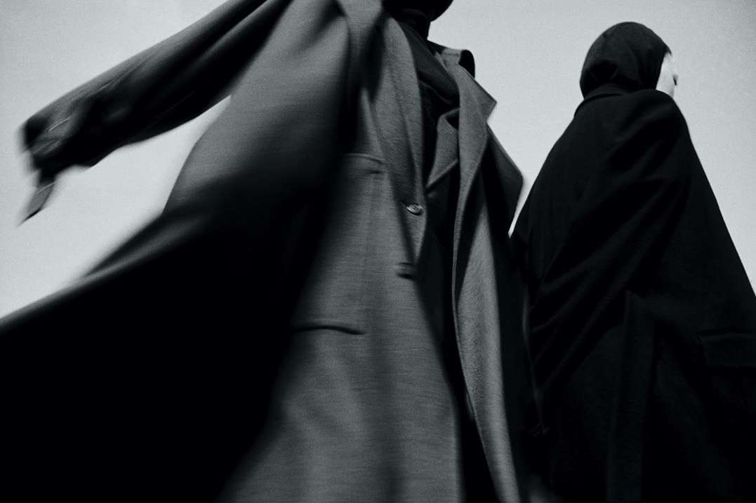 Two figures in draped coats against a bright background showcase dark, minimalist fashion with subtle motion blur and stark contrast.