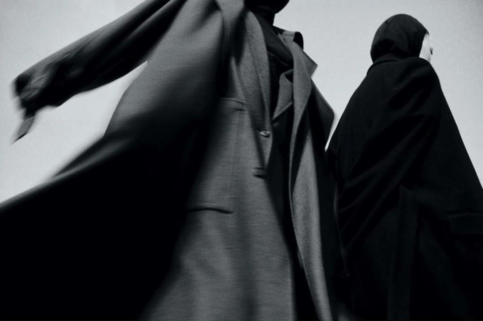 Two figures in draped coats against a bright background showcase dark, minimalist fashion with subtle motion blur and stark contrast.