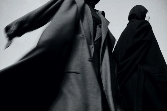 Two figures in draped coats against a bright background showcase dark, minimalist fashion with subtle motion blur and stark contrast.