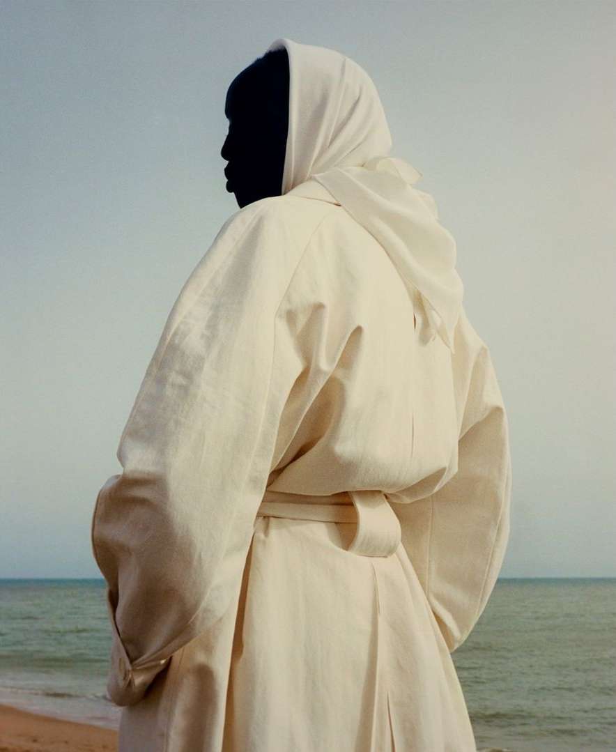 Woman in white coat and headscarf stands on a beach with a muted blue sea and sky creating a minimalist, serene outdoor fashion scene.