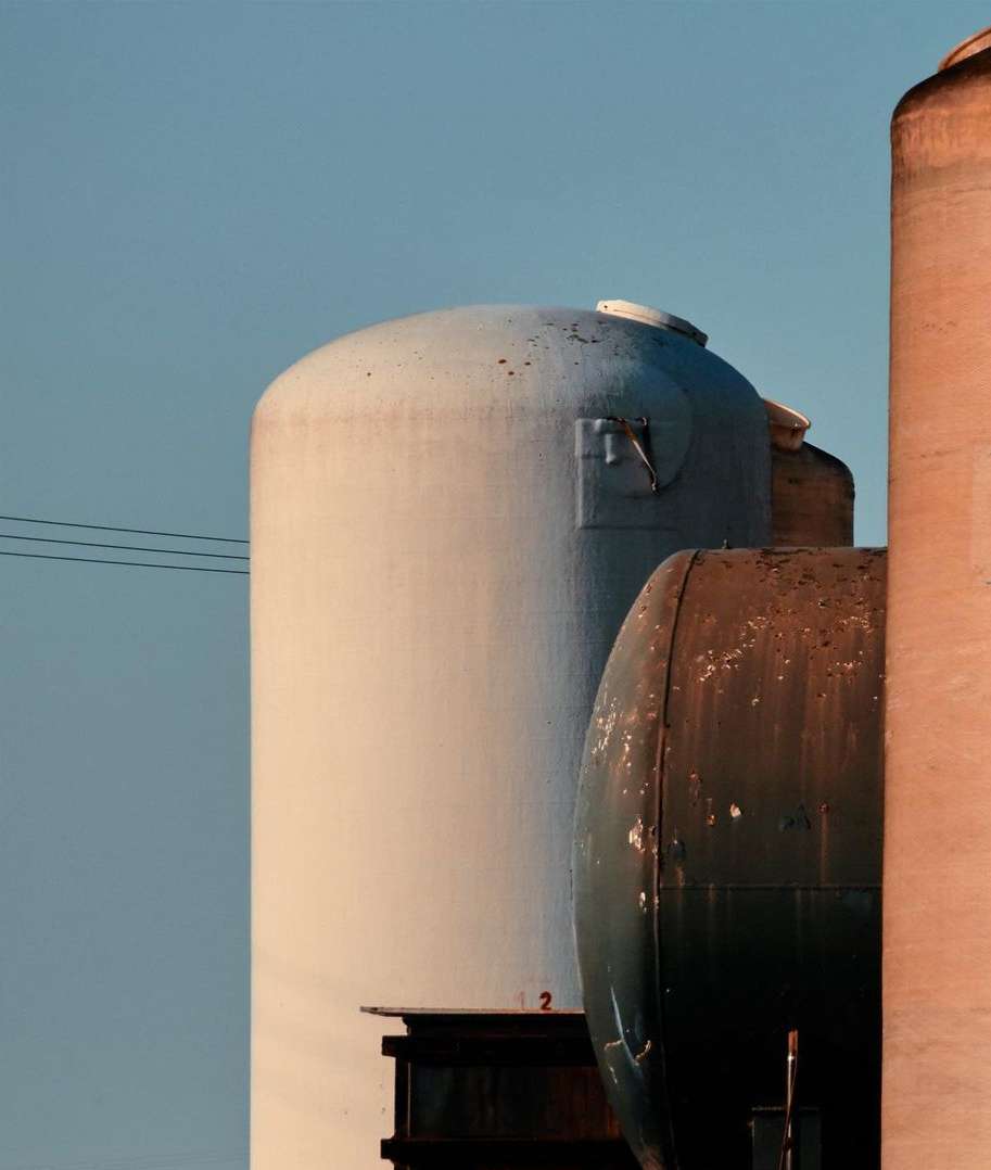 Industrial storage silos stand against a clear sky, featuring a white, cylindrical tank and a dark, rounded tank, with power lines overhead.