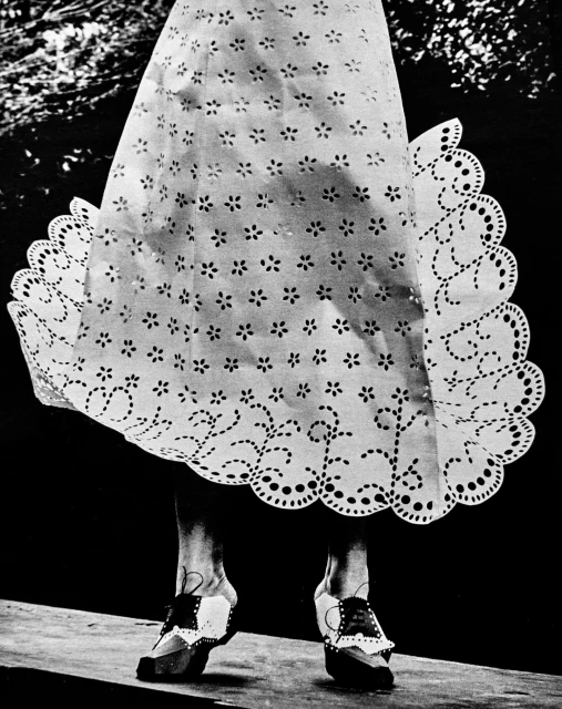 Woman wearing a white eyelet lace dress and unique shoes standing on a light-colored surface against a dark background with foliage.