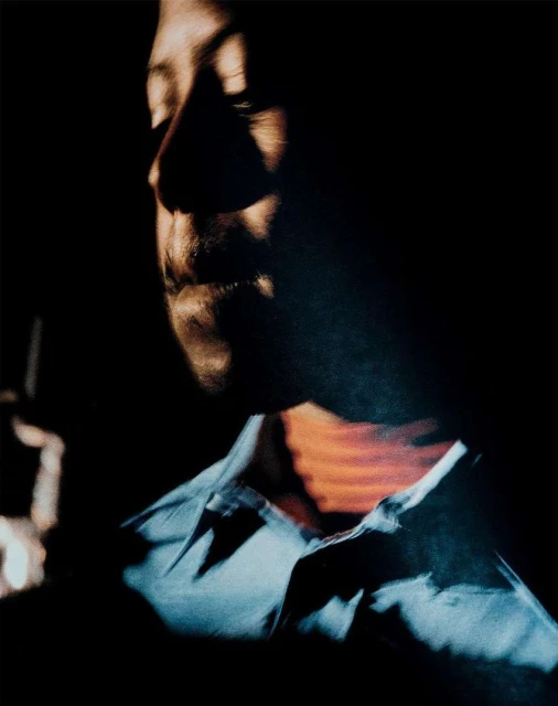 Low-lit portrait of an adult with a light blue shirt. The composition features dark shadows and minimal background detail.