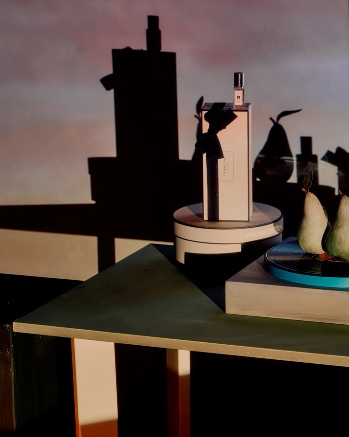Still life with perfume bottle and pears on a geometric table, with shadows casting against a light pink and blue background.