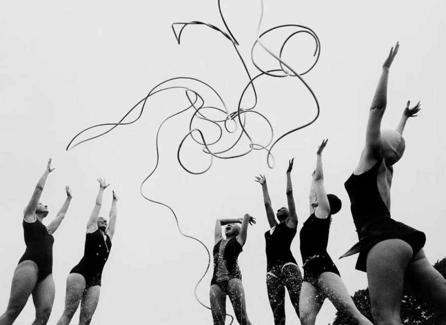 A group of six women in swimsuits with their arms raised towards a tangled wire formation against a bright sky backdrop.