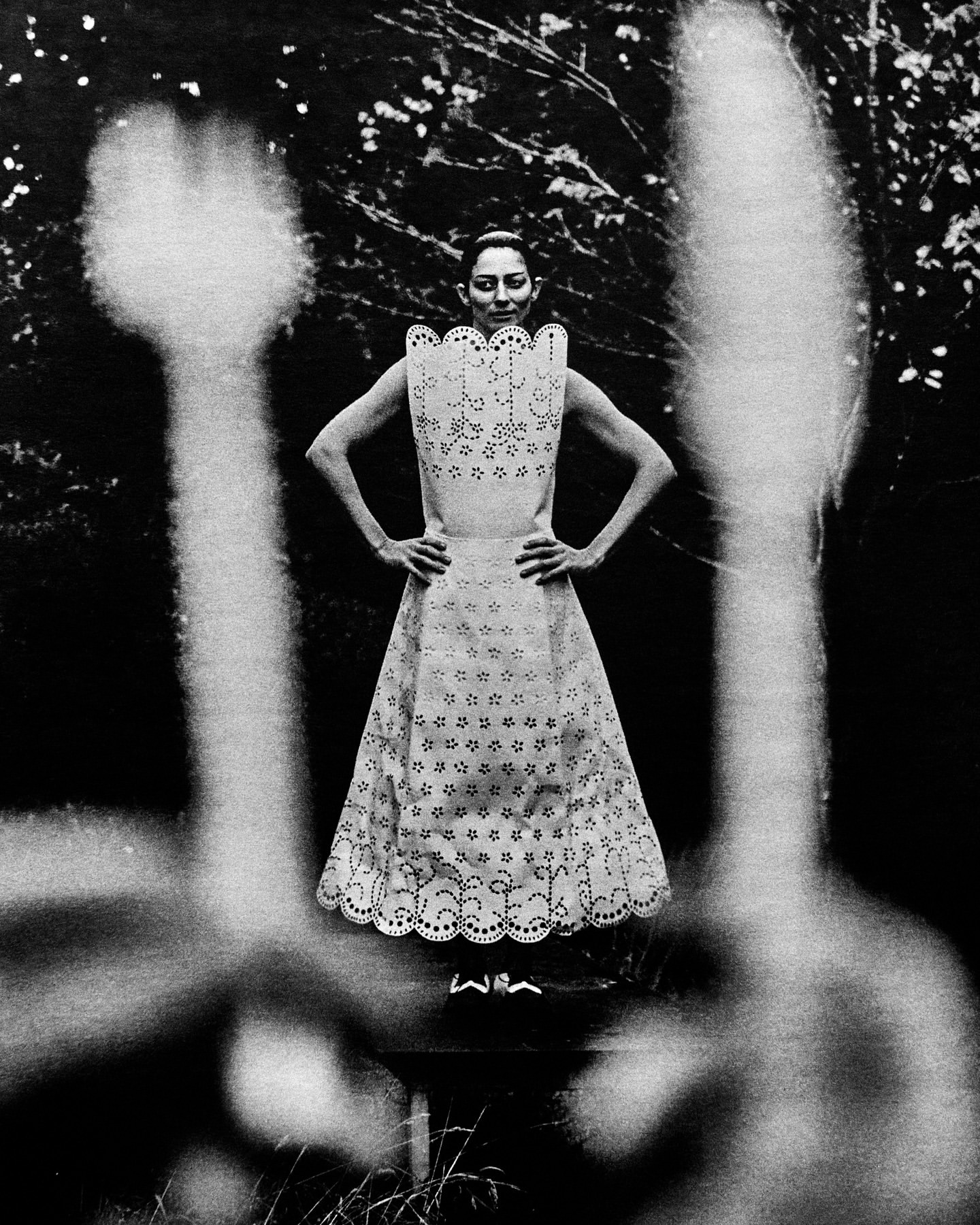 Monochrome portrait of a poised woman in an eyelet dress, framed by blurred flora against a backdrop of foliage and natural light.