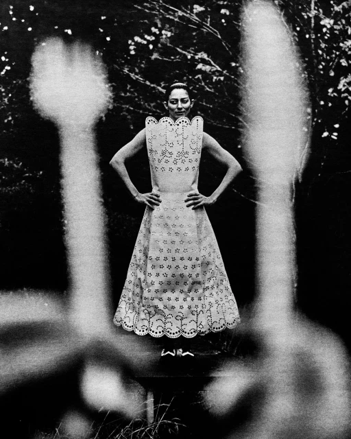 Monochrome portrait of a poised woman in an eyelet dress, framed by blurred flora against a backdrop of foliage and natural light.