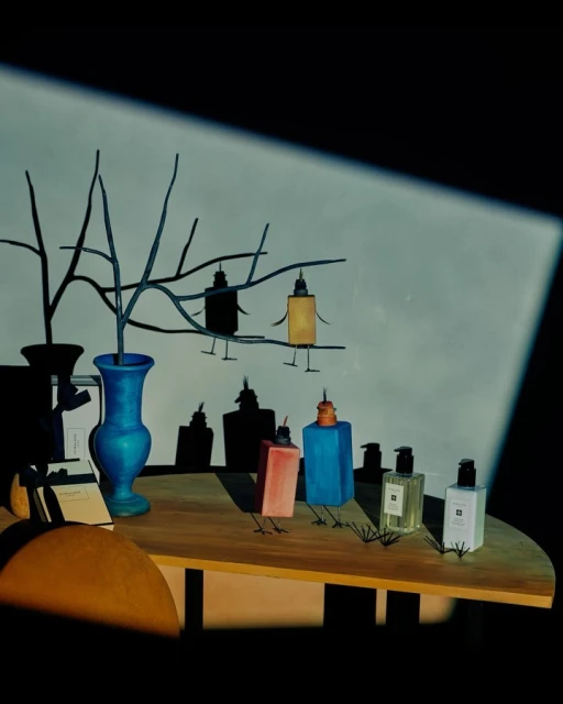 Whimsical still life featuring perfume bottles with added legs, arranged on a table with a vase and branch in a brightly lit, indoor setting.