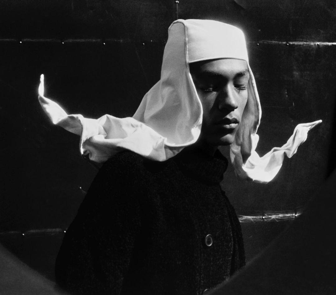 Portrait of a person with downcast eyes wearing a white bonnet and a dark coat, captured in a black-and-white studio setting.