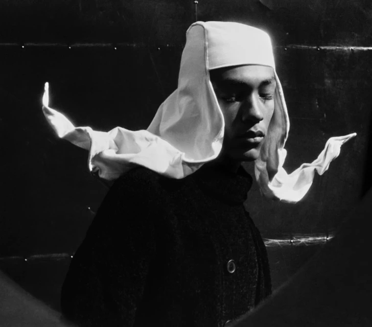 Portrait of a person with downcast eyes wearing a white bonnet and a dark coat, captured in a black-and-white studio setting.