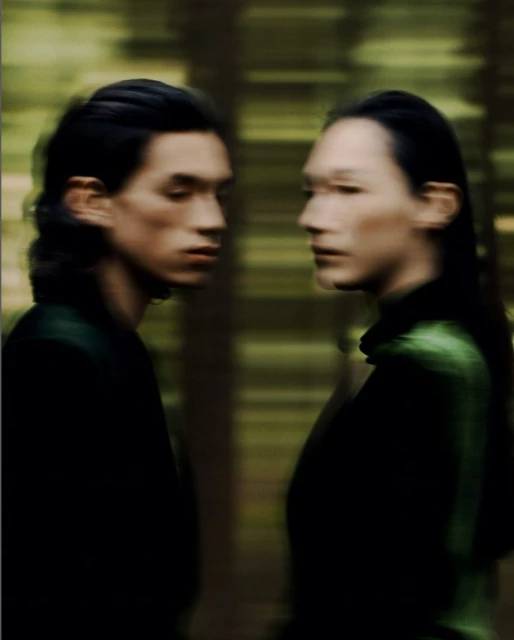 Two people stand in a blurred forest setting, facing each other, illuminated with natural light, wearing dark clothing in a medium shot.