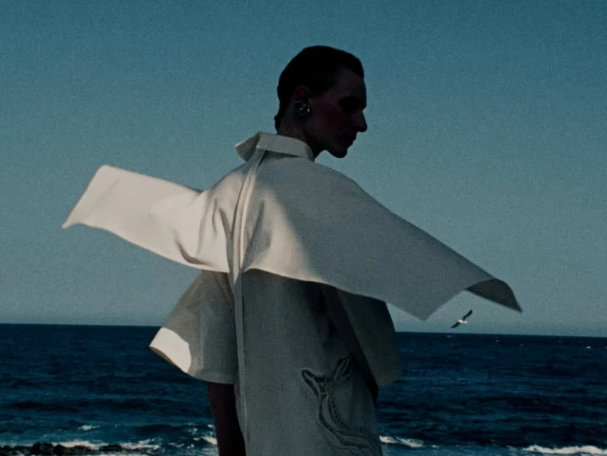 Adult fashion model in light-colored, oversized coat posing on rocky coastline with turquoise ocean and clear blue sky in outdoor setting.