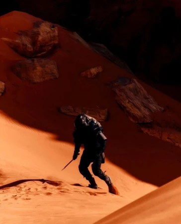Person in dark tactical clothing holding blade, walking on sand dune with rocks in orange-lit desert environment.