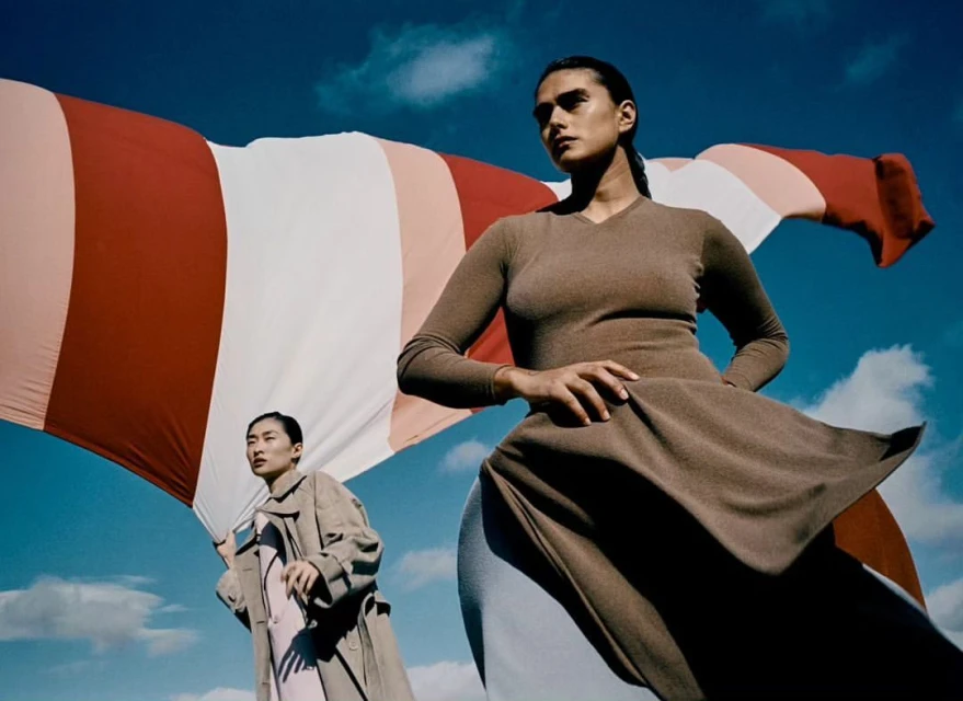 Two women pose outdoors against a backdrop of sky and a large striped sheet, exuding editorial fashion and a serene yet confident mood.