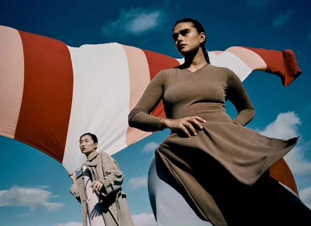 Two women pose outdoors against a backdrop of sky and a large striped sheet, exuding editorial fashion and a serene yet confident mood.