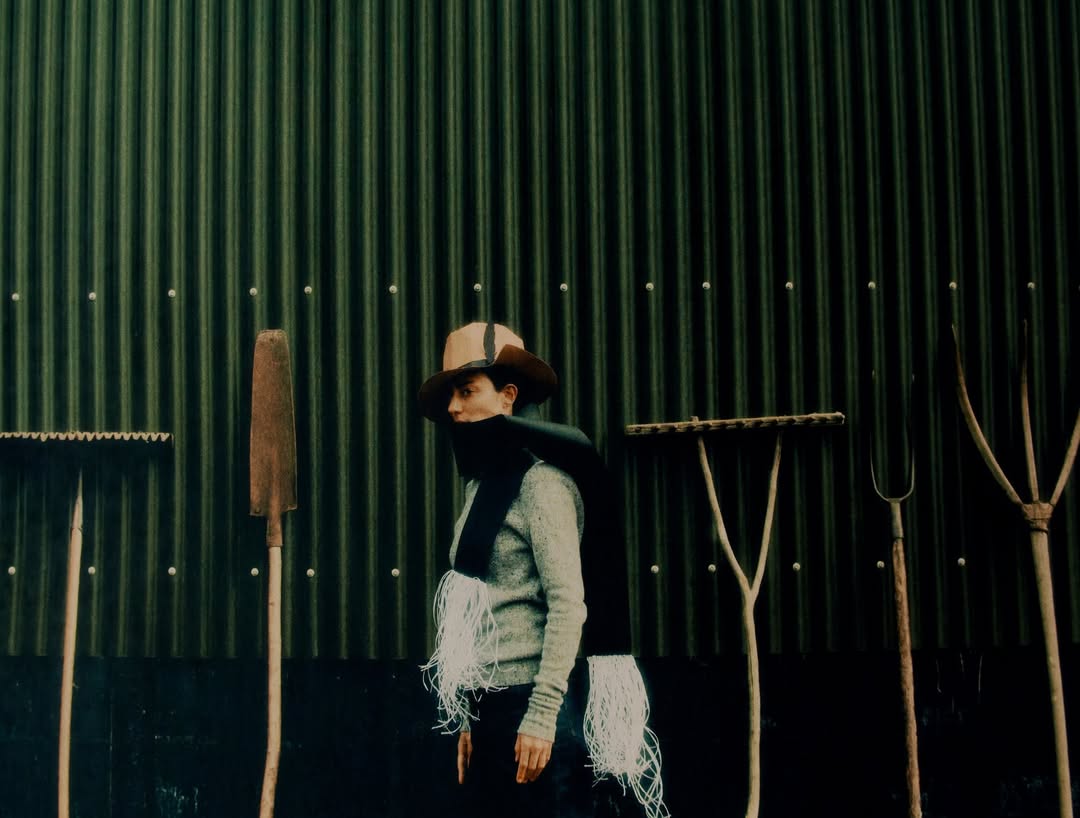 Person in a hat and scarf poses in front of a green corrugated metal wall with various garden tools arranged on the background.