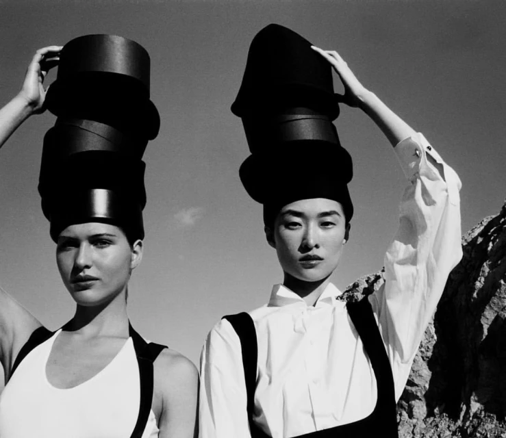 Two fashion models in white tops and black bottoms pose outdoors with oversized black hats, set against a bright, sunny sky.