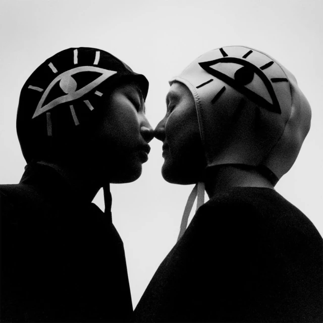 Close-up black and white portrait of two women in swim caps with eye designs, faces touching, set against a stark white background.