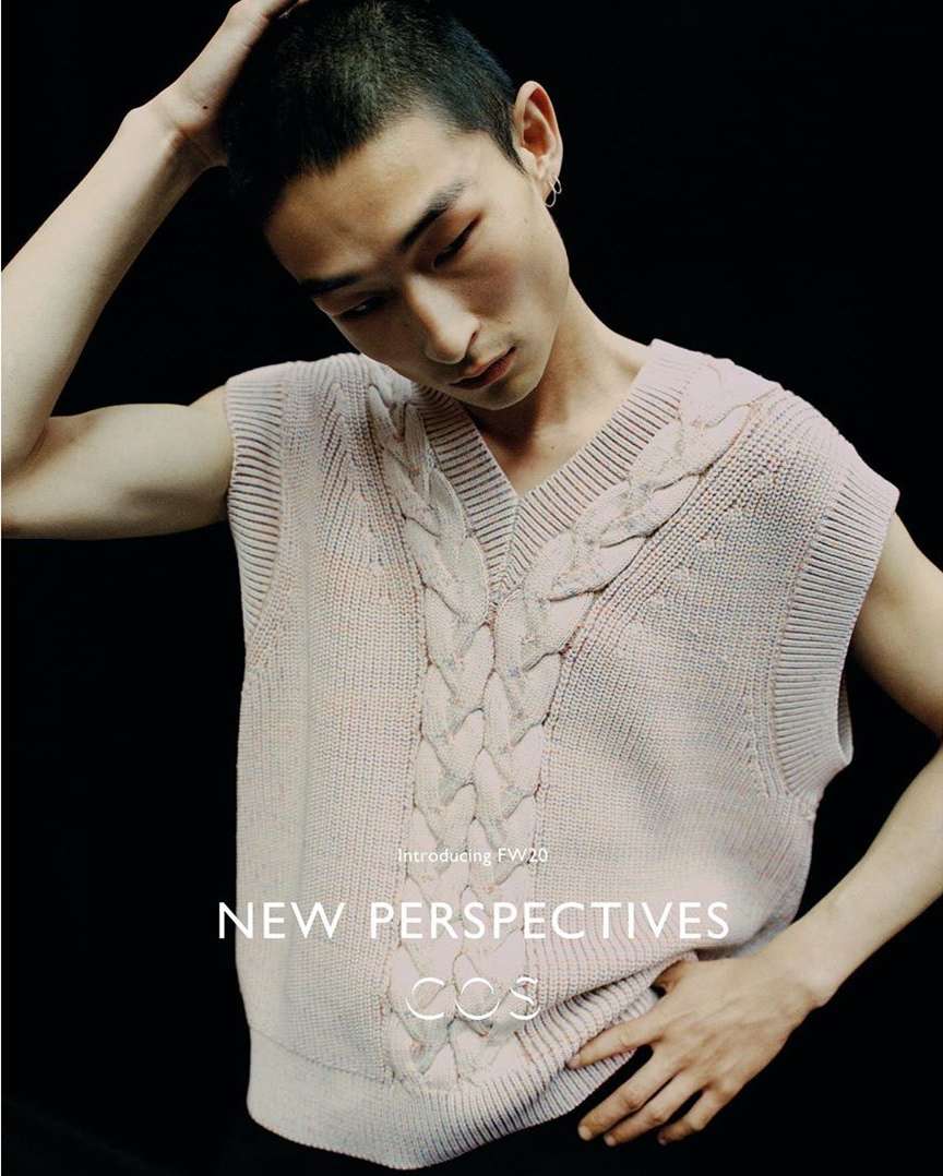 Close-up of an adult man wearing a cream knitted sweater vest with a V-neck, posing against a dark background in a fashion setting.