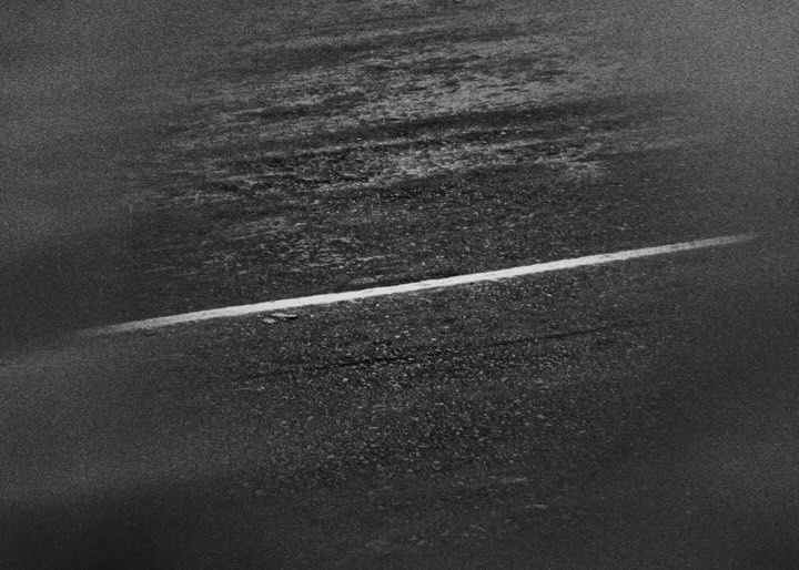 Monochrome shot of an asphalt road with a single white lane marking, featuring a wet, reflective surface and grainy texture.