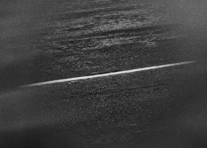 Monochrome shot of an asphalt road with a single white lane marking, featuring a wet, reflective surface and grainy texture.