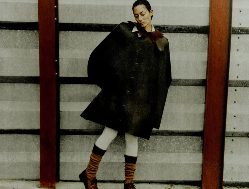 A stylish woman poses outdoors wearing a dark cape, white tights, brown knee socks, and loafers, against a gray and red background.