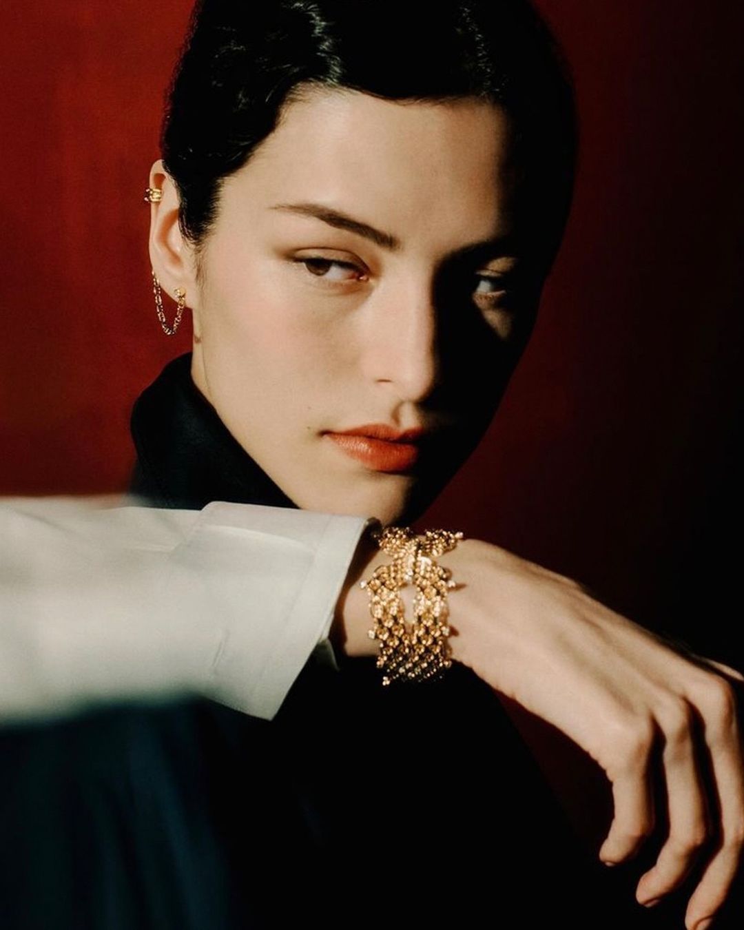 Portrait of a woman with dark hair in a black top and white sleeve, wearing a gold bracelet, against a dark red background.