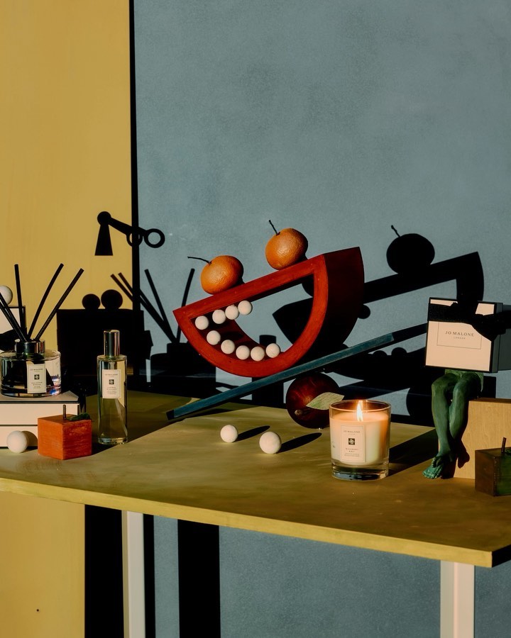 Still life with fragrance bottles, candle, decorative shapes, and oranges on a tabletop against a split yellow and blue wall.