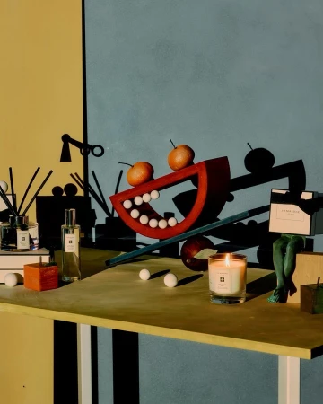 Still life with fragrance bottles, candle, decorative shapes, and oranges on a tabletop against a split yellow and blue wall.