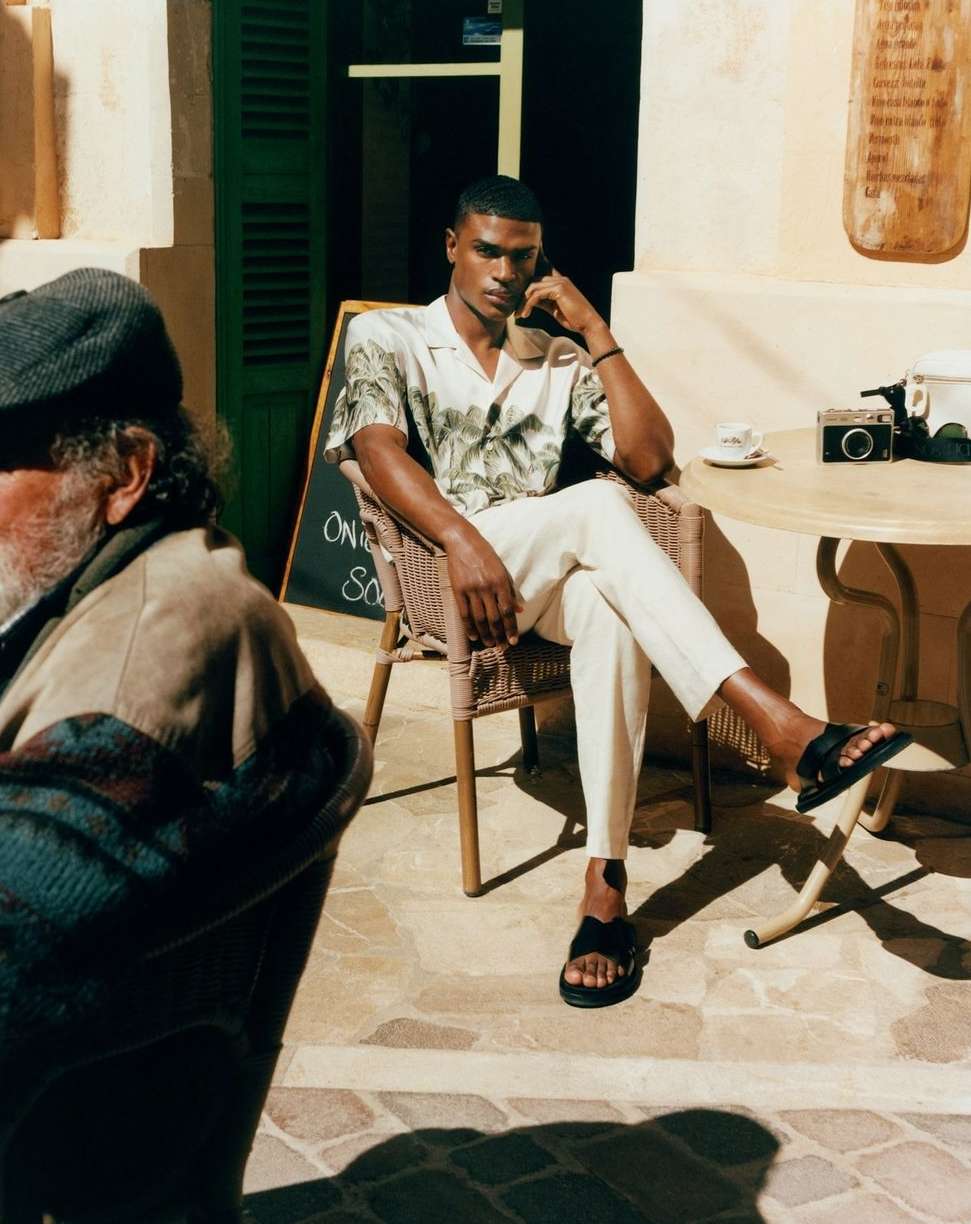 A dark-skinned male model sits outside a café, wearing a patterned shirt, black sandals, and white pants, captured in soft, diffused light.